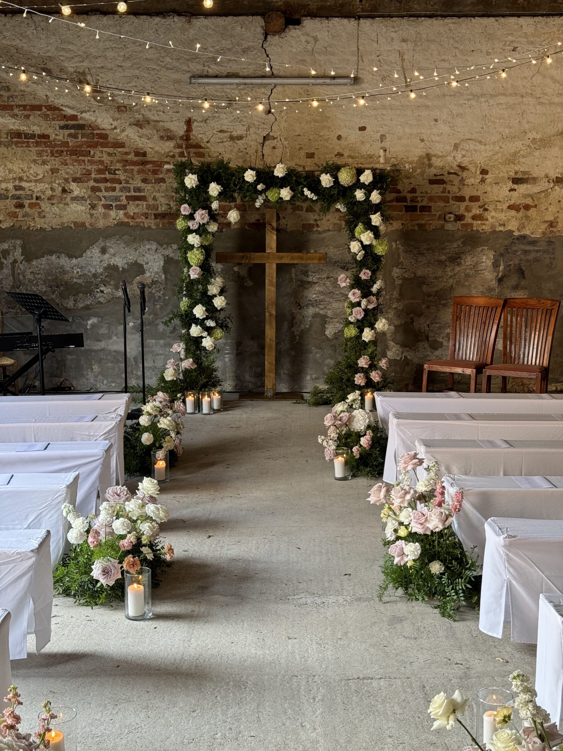 Eckiger Blumenbogen vor dem Kreuz einer altertümlichen Kirche hinter weißen Bänken und hellen, großen Blumensträußen
