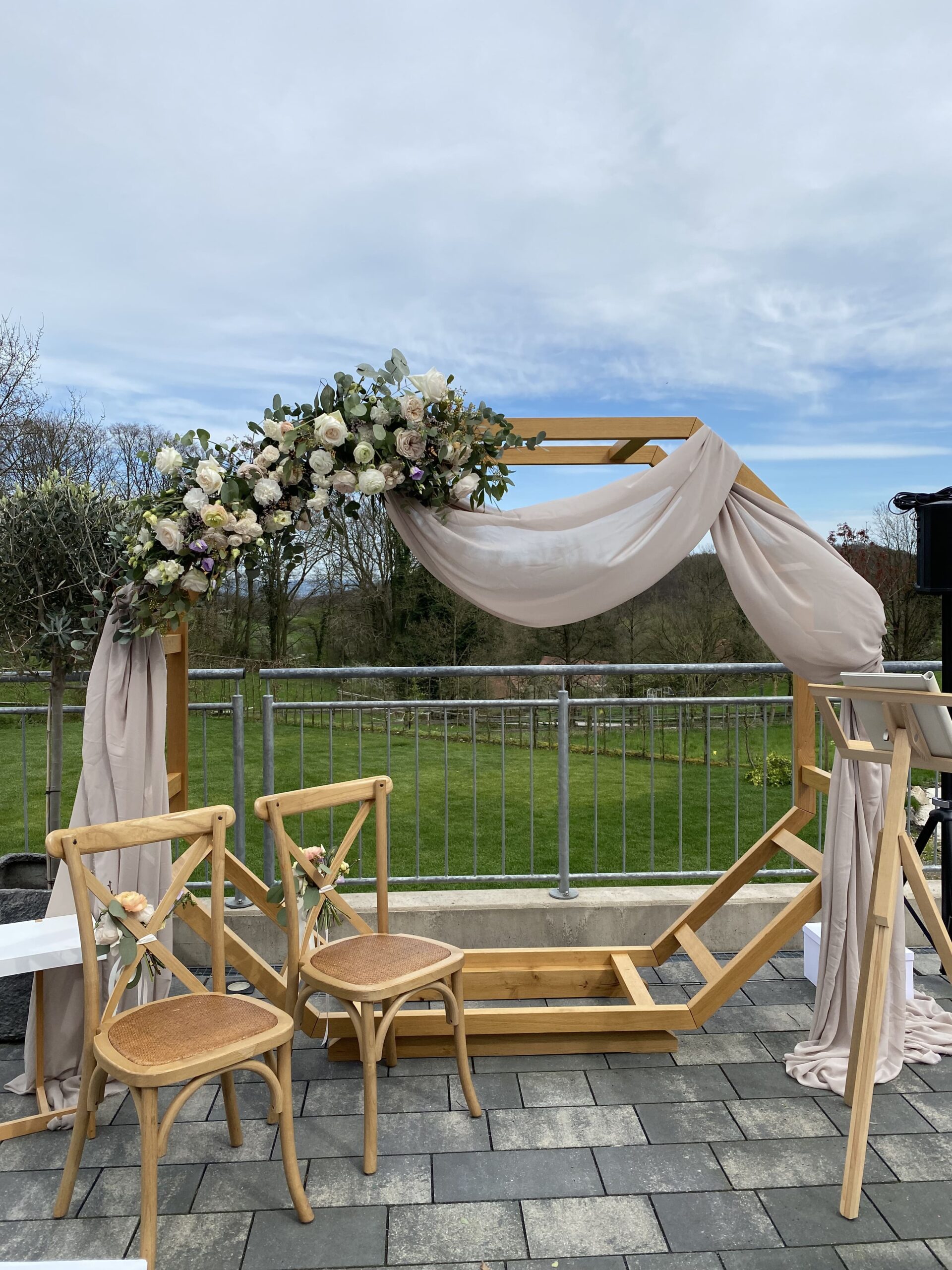 Großer Blumenbogen aus Holz mit einem beigen Schleier hinter zwei Stühlen aus Echtholz auf einer Außenterrasse