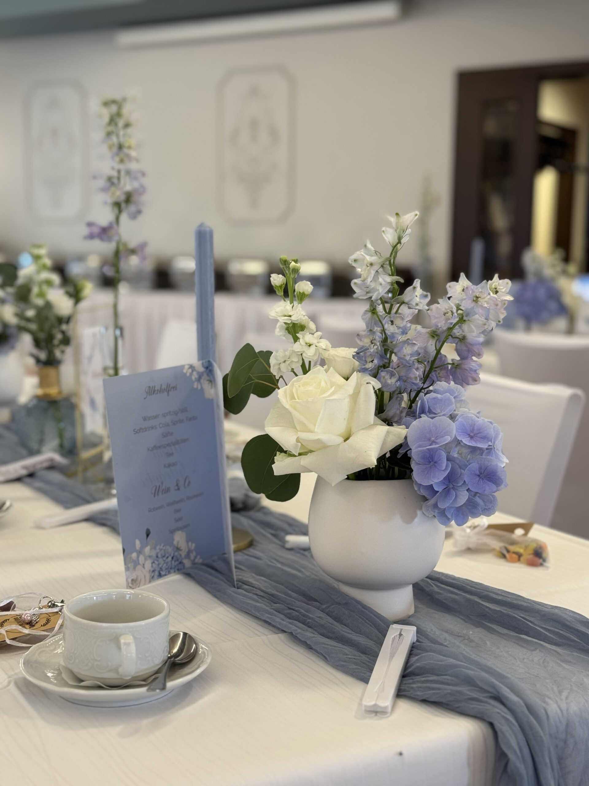 Blumen in weiß und blau in einer weißen Vase auf einem gedeckten Tisch