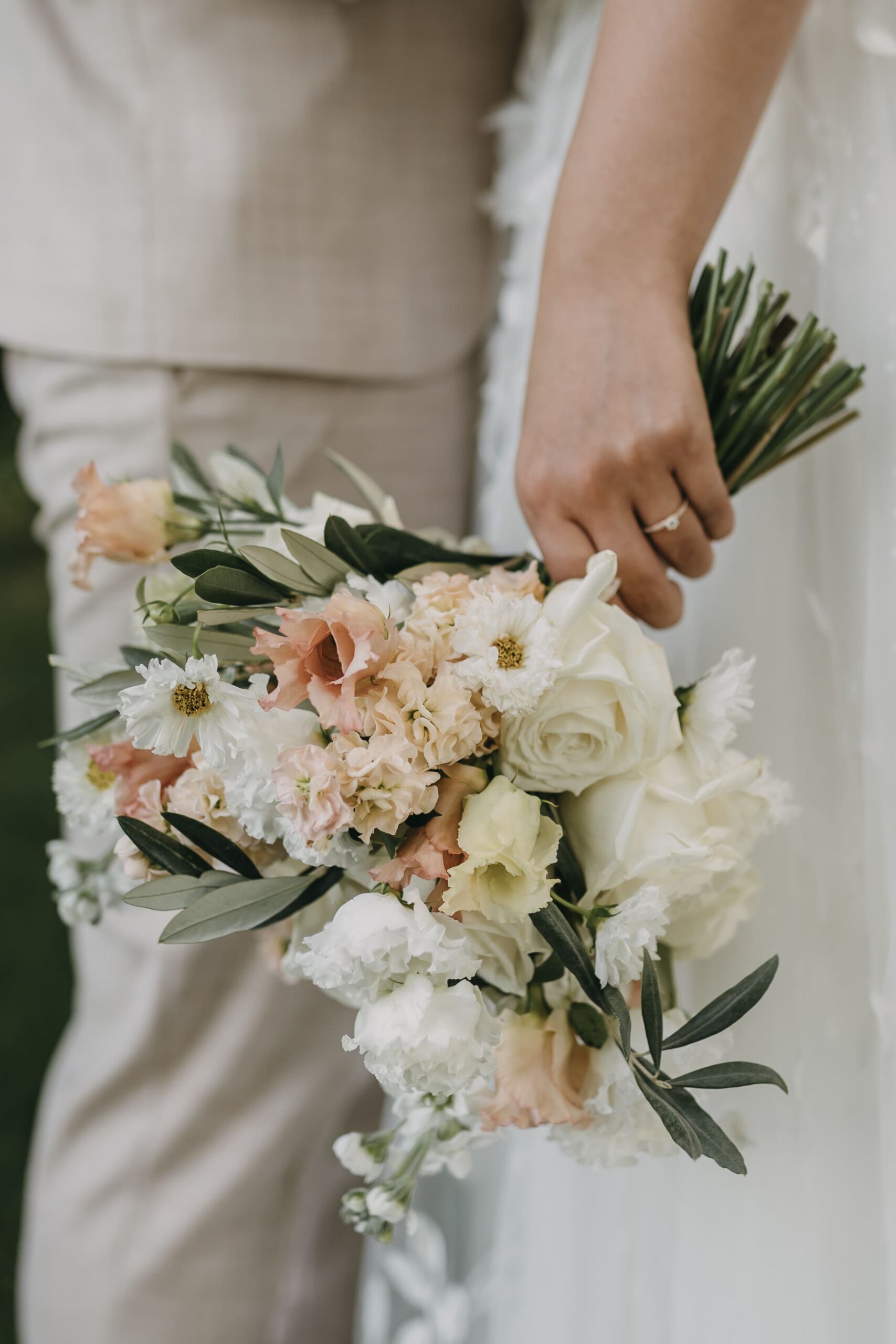 Die linke Hand der Braut mit Ring am Ringfinger und Brautstrauß in der Hand mit hellen Blumen