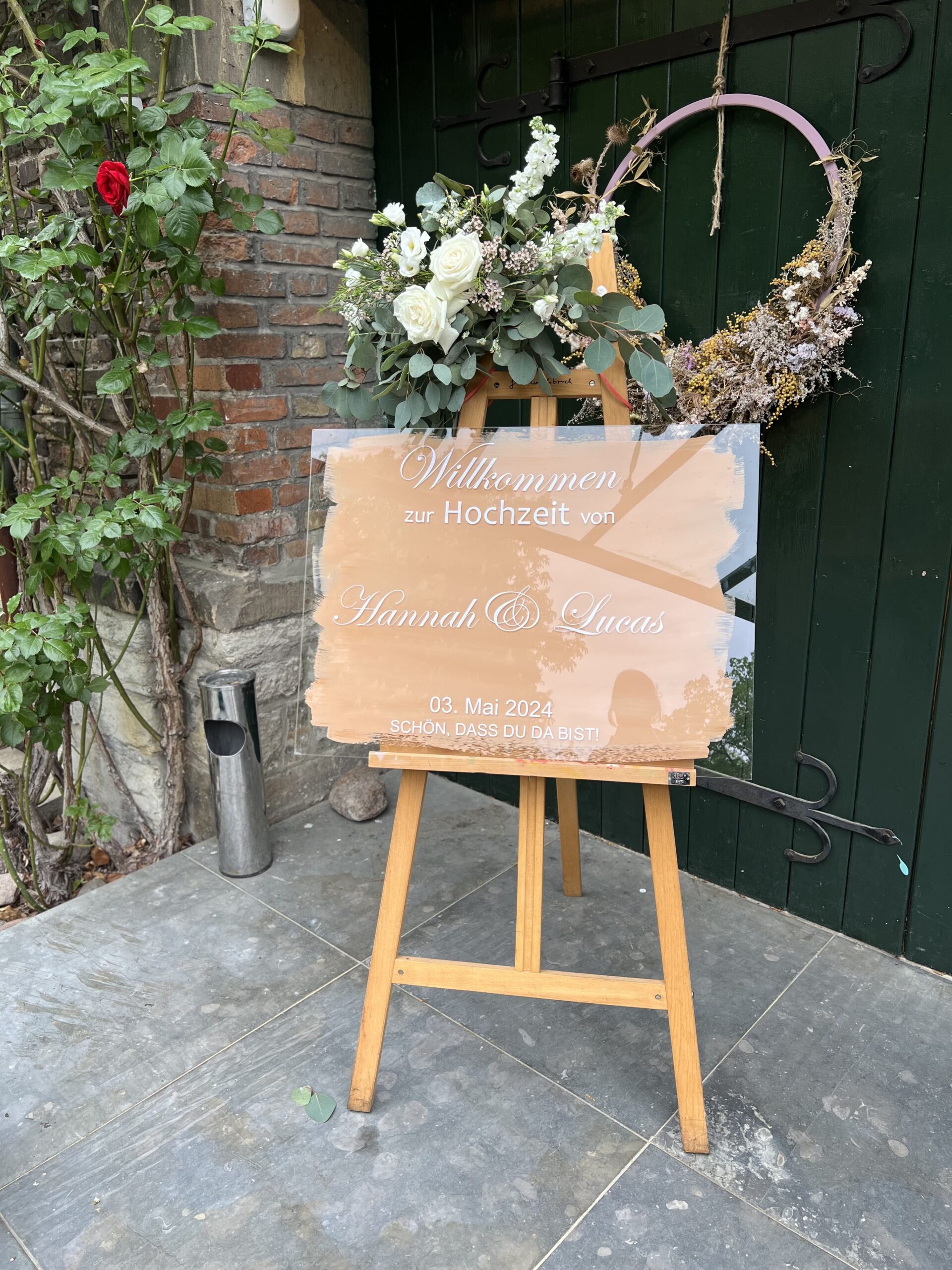 Ein Willkommensschild aus Glas zur Hochzeit vor einem Eingang mit Blumen und Grün