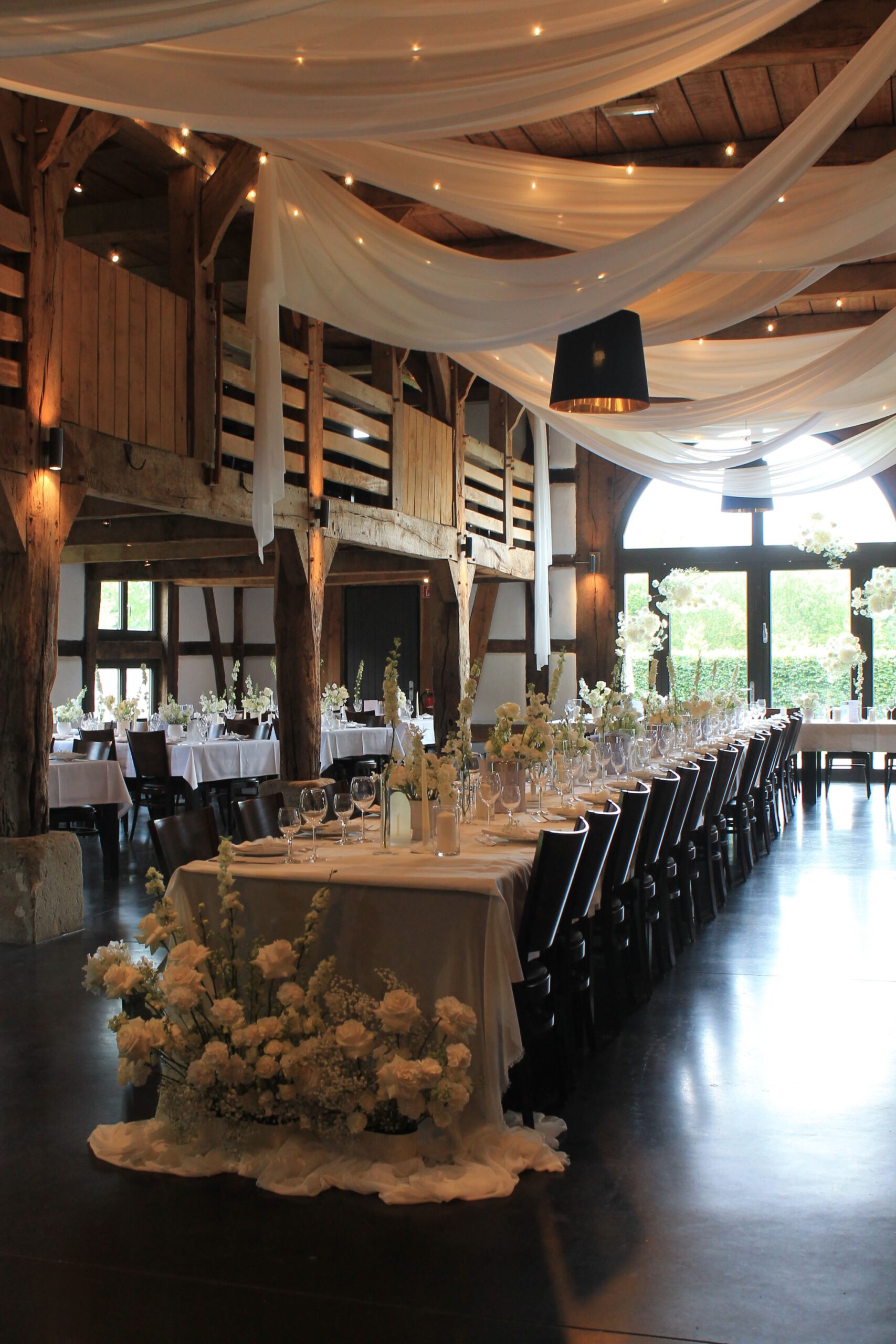 Ein langer Gästetisch mit vielen weiße, großen Rosen am Tischanfang und weiße Schleier über dem Tisch hängend