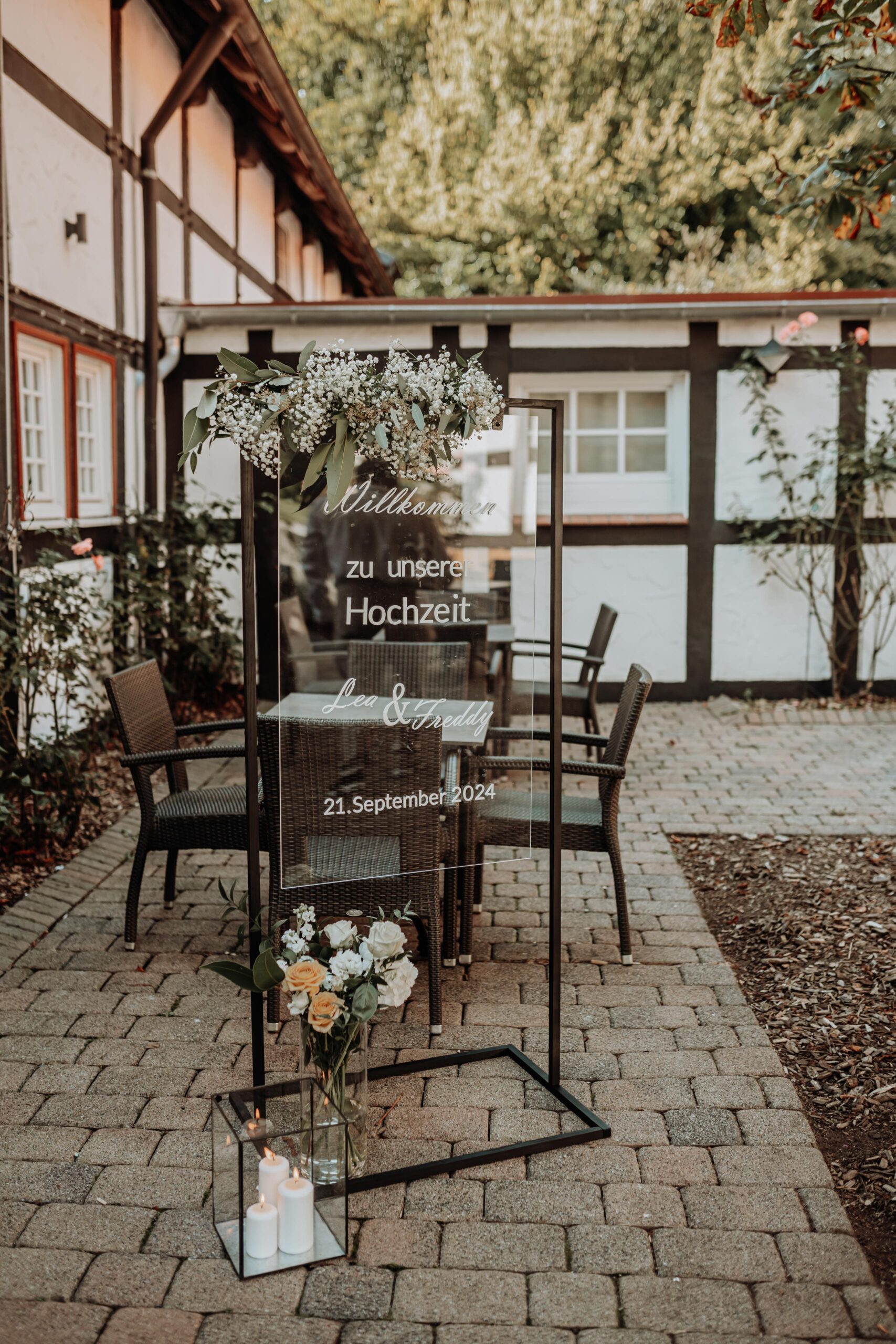 Willkommensschild aus Glas mit weißer Schrift die die Gäste vor einem Fachwerkhaus zur Hochzeit begrüßt